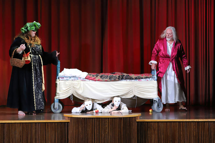 Teaterscen, kvinna och man med en säng i mitten. Masker under sängen.