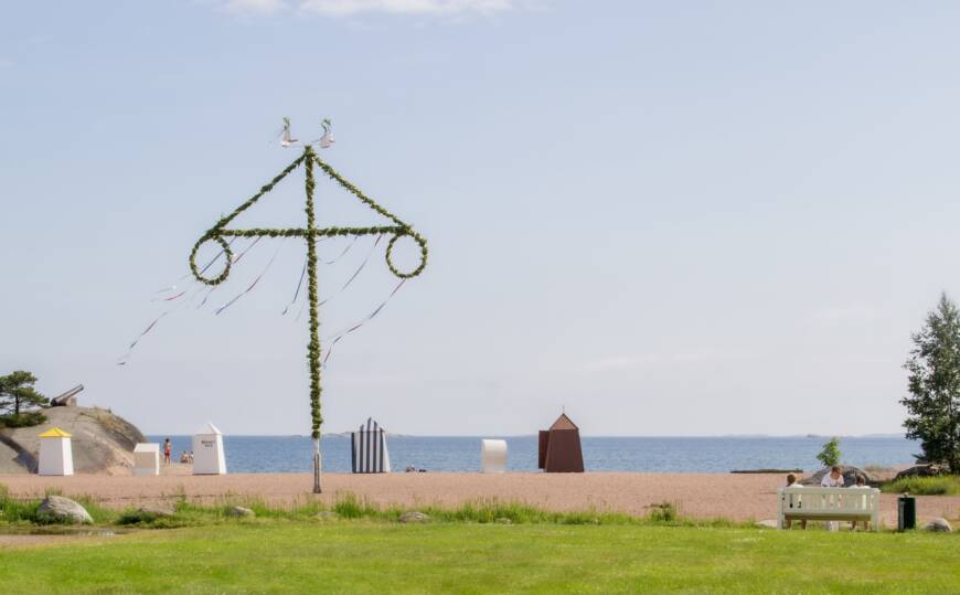 En solig sommardag med vy över midsommarstången, strand och grön gräsmatta.