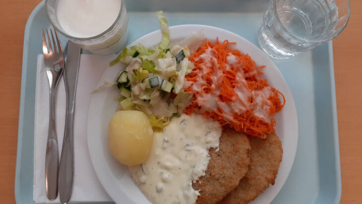 Tarjottimella lautanen, jossa peruna, salaattia, porkkanaa, kastiketta ja pihvejä. Kaksi lasia ja aterimet.