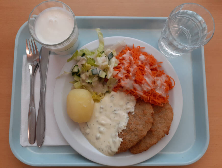 Tarjottimella lautanen, jossa peruna, salaattia, porkkanaa, kastiketta ja pihvejä. Kaksi lasia ja aterimet.